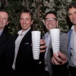 Cuatro hombres trajeados están de pie al aire libre por la noche, dos de ellos levantan hacia la cámara vasos reutilizables blancos y estriados. En el fondo hay plantas y luces, lo que sugiere un evento festivo o formal. -corvt_ar