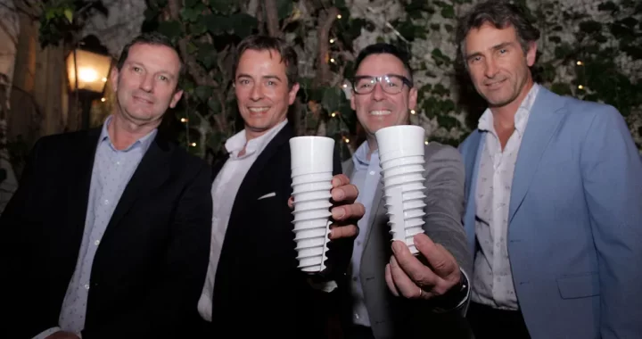 Cuatro hombres trajeados están de pie al aire libre por la noche, dos de ellos levantan hacia la cámara vasos reutilizables blancos y estriados. En el fondo hay plantas y luces, lo que sugiere un evento festivo o formal. -corvt_ar