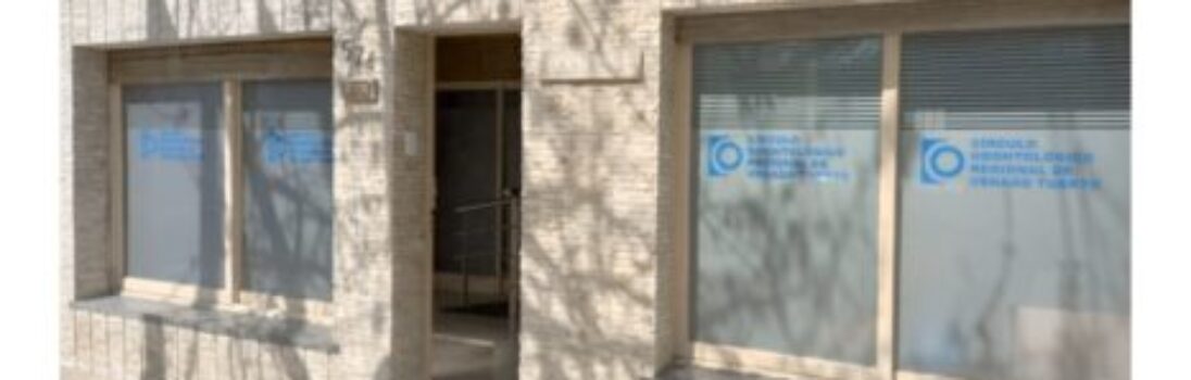 Entrada de un edificio de ladrillo beige con dos grandes ventanas que muestran texto y logotipos azules de Odontotal Circulo Odontologico, parcialmente cubierto por sombras de ramas de árboles. La puerta está abierta y deja ver un pasillo interior. -corvt_ar